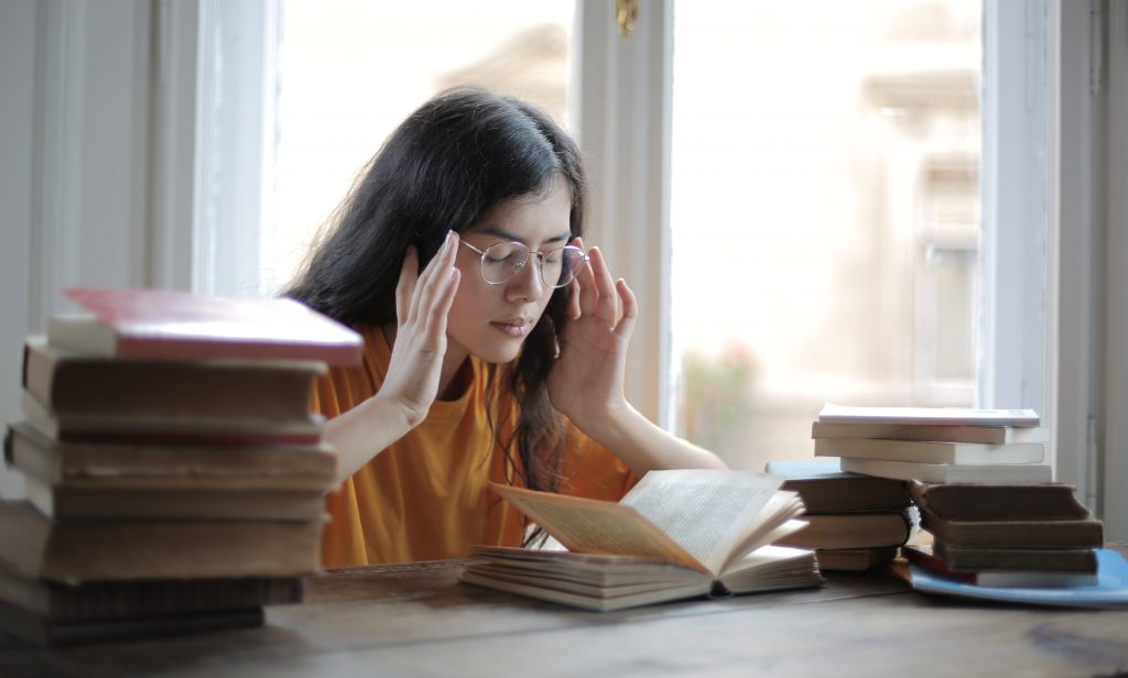 Eine junge Frau sitzt neben mehreren Bücherstapeln. Sie richtete ihre Brille mit den Händen seitlich während sie ein aufgeschlagenes Buch vor sich liest.