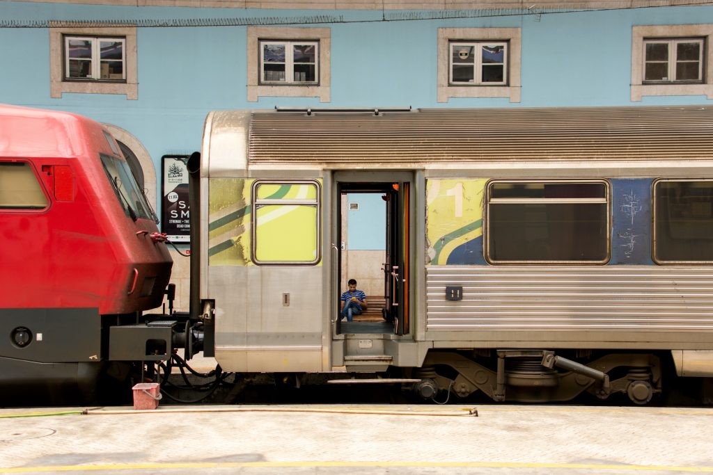 Ein Zug mit roter Lok steht im Bahnhof und hat die Türen offen stehen. Man sieht eine Person auf einer Bank dahinter warten. Das Bild soll hier "Verspätung" darstellen.