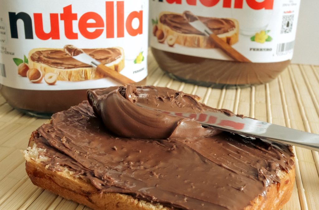 Ein Brot mit Nutella-Aufstrich liegt auf einem weißen Teller vor zwei Gläsern Nutella.