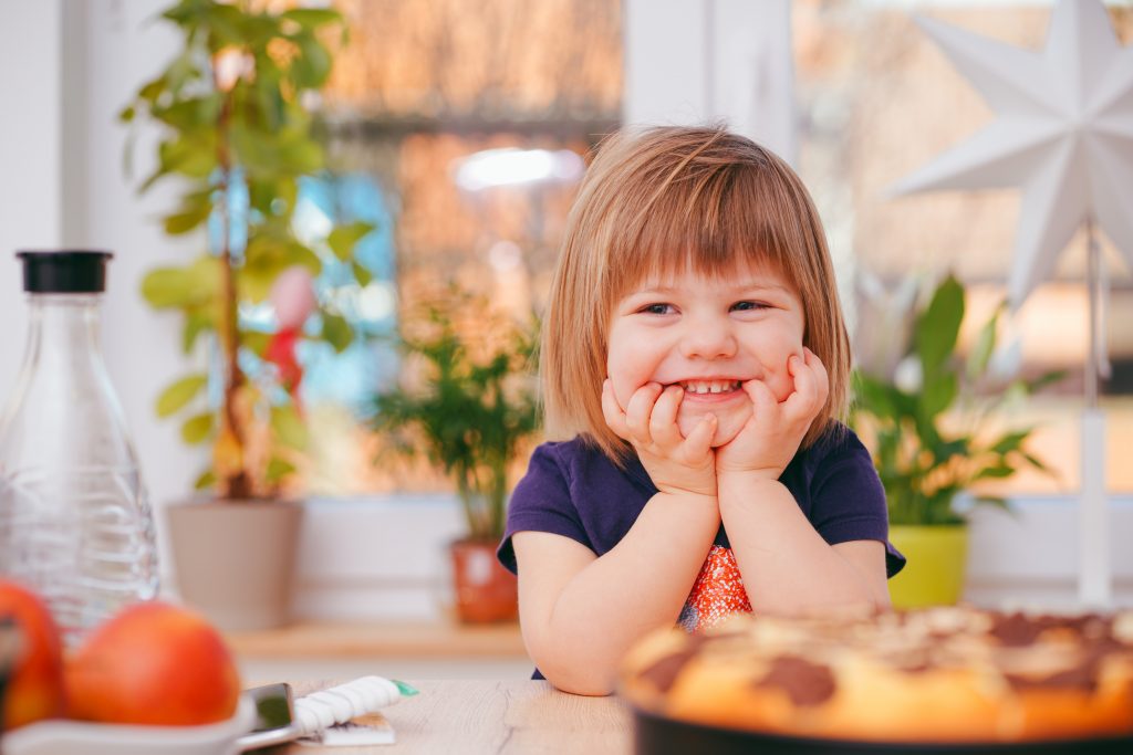 Ein kleines, blondes Mädchen sitzt an einem Holztisch auf dem Essen steht und lacht. Es hält die Hände am Kinn.