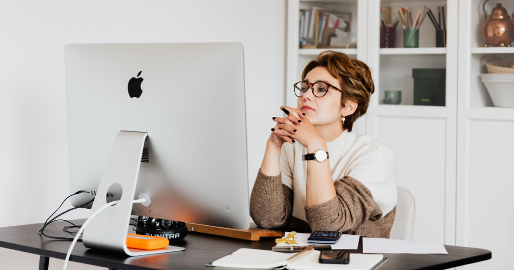 Eine Frau sitzt sichtlich genervt vor einem Computerbildschirm mit vielen Papieren neben ihr auf einem Schreibtisch. Gehört zu einem Blog über Outlook und Mailprogramme.