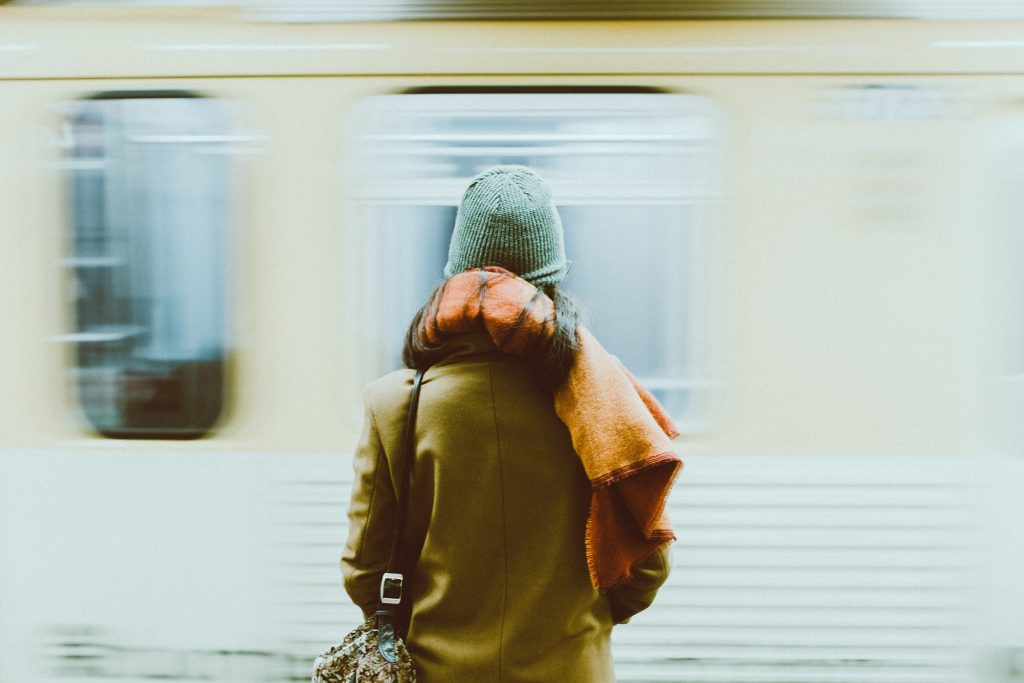 Eine Frau in brauner Jacke steht an einem Bahnsteig, an dem mit hoher Geschwindigkeit ein Zug vorbeifährt.
